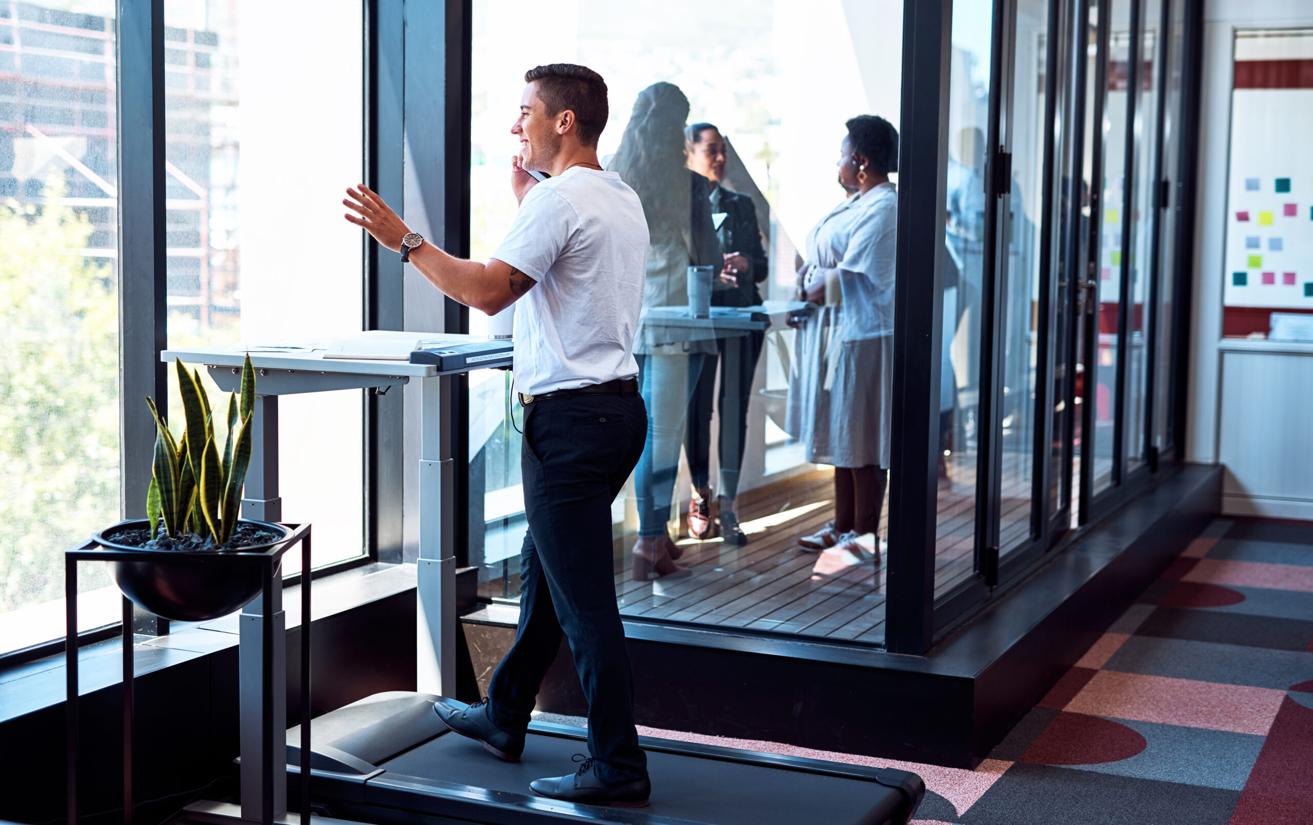 Cette image illustre un environnement de travail moderne et dynamique où un homme utilise un tapis de course avec un bureau debout, tout en prenant un appel téléphonique. La scène montre un espace ouvert avec des grandes fenêtres, apportant beaucoup de lumière naturelle. À l'arrière-plan, on peut voir d'autres collègues engagés dans des discussions près d'autres bureaux debout, ce qui reflète une atmosphère de travail collaborative et active. Le fait de marcher tout en travaillant, comme le montre cet homme, met en évidence l'intégration de l'exercice dans la routine professionnelle pour promouvoir la santé et la productivité. Les plantes en pot ajoutent une touche de verdure à l'espace, contribuant à un environnement de travail agréable et stimulant.