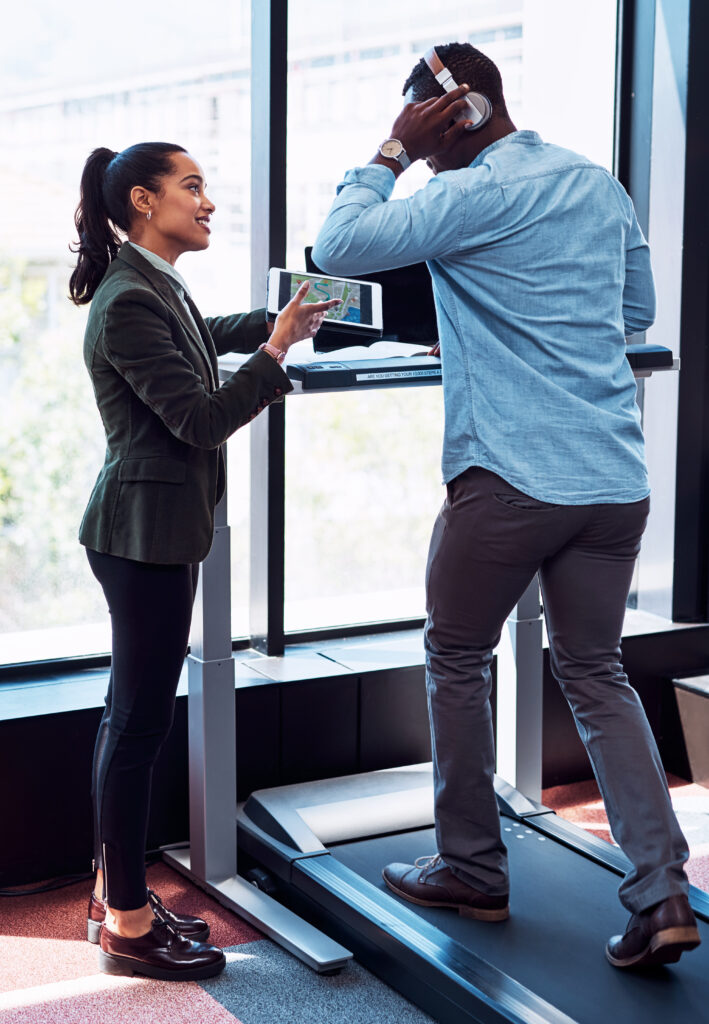 Un professionnel utilise un tapis de marche bureau tout en discutant avec une collègue, combinant activité physique et collaboration au travail. Un exemple de l'intégration harmonieuse de la santé et de la productivité dans l'environnement de bureau moderne.