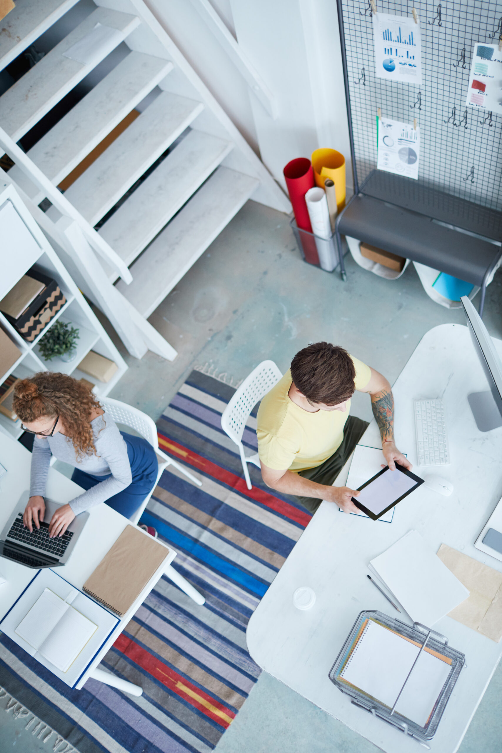 Vue aérienne d'un espace de travail moderne et créatif, mettant en scène deux professionnels concentrés sur leurs tâches respectives. Un environnement lumineux et bien organisé, idéal pour stimuler la productivité et la collaboration.